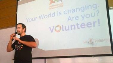 A man wearing a black t-shirt standing and speaking into a microphone. The background, there is a screen with the words "Your world is changing - are you? Volunteer!" projected behind him.