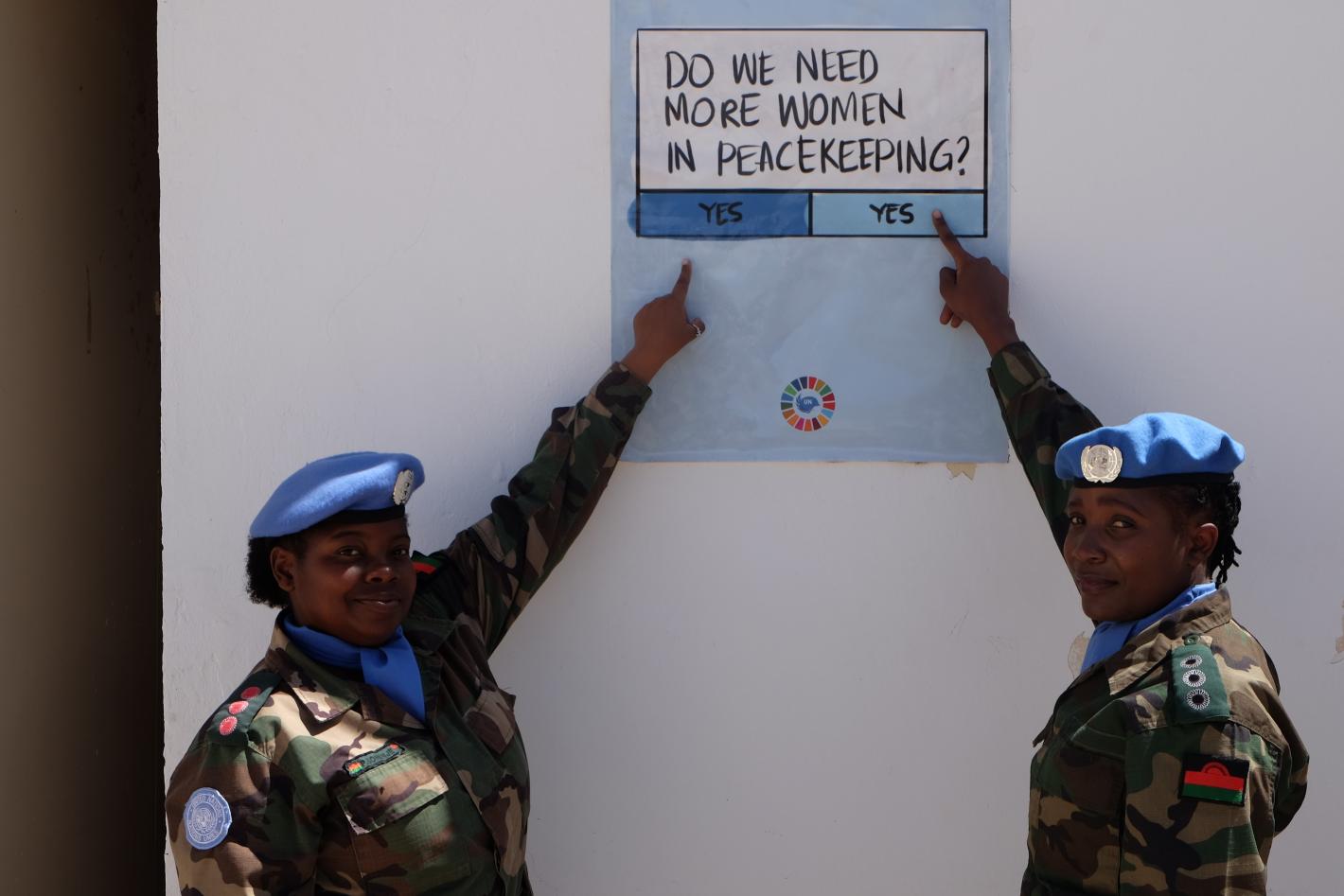 Two women peacekeepers in military uniform point at a sign that poses the question "do we need more women in peacekeeping" and gives "yes" or "yes" as possible responses. 