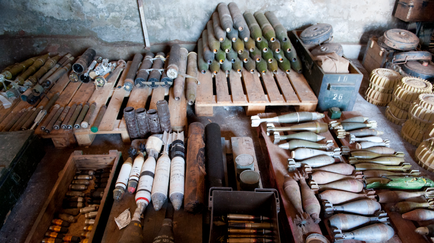 Various types of ammunition and explosives, including mortar and artillery shells, are organized on pallets and crates in a storage area.