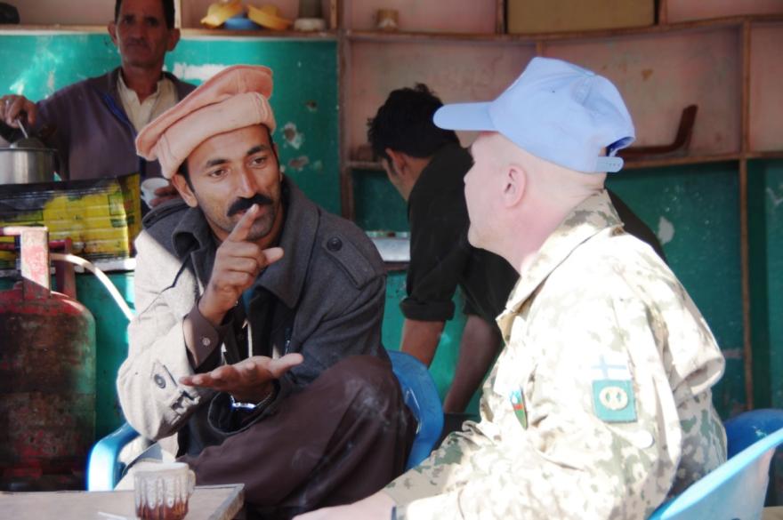 UN peacekeeper in camouflage uniform and blue beret sits indoors, talking with a local person wearing traditional attire. A tea glass and kitchen items are visible.
