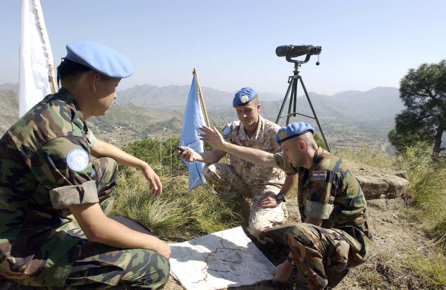 Three UN peacekeepers in blue helmets review a map on a hillside with binoculars mounted on a tripod nearby, overlooking a valley with mountains in the background.