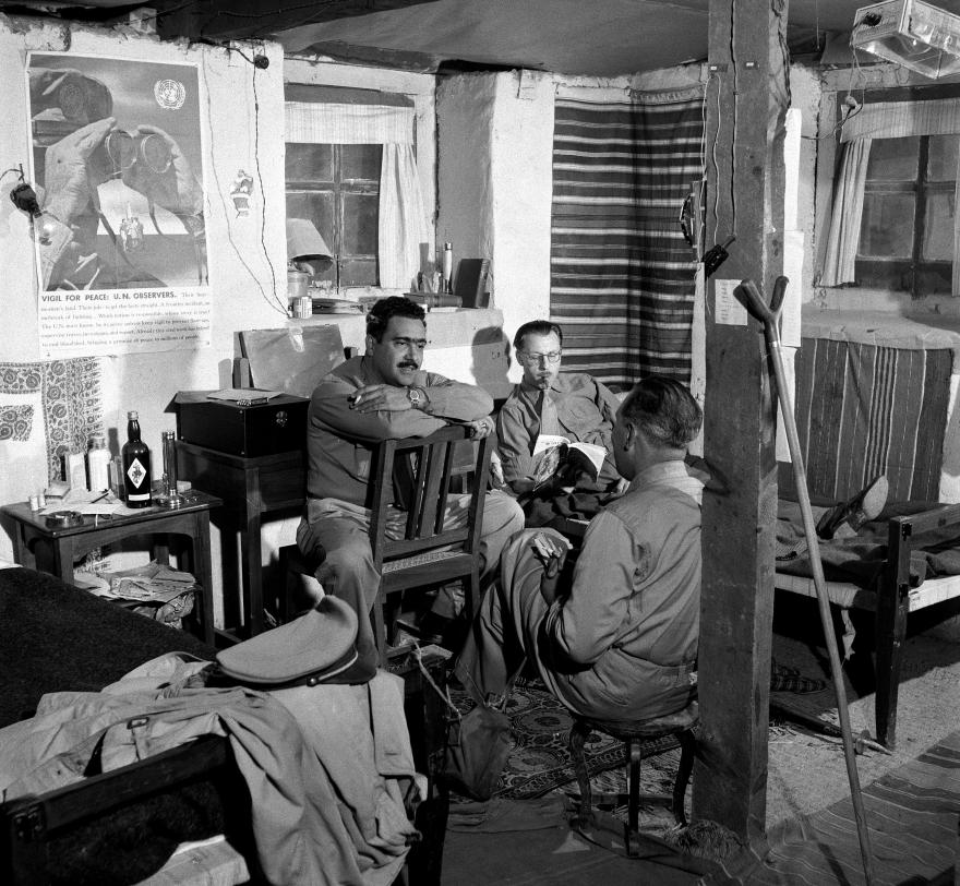 Three UN military observers sit inside a modest living quarters with simple furnishings, including a bed, wooden chairs, and a table with bottles. A poster on the wall reads “Vote for Peace! U.N. Observers.” The room has patterned textiles and a window with curtains.